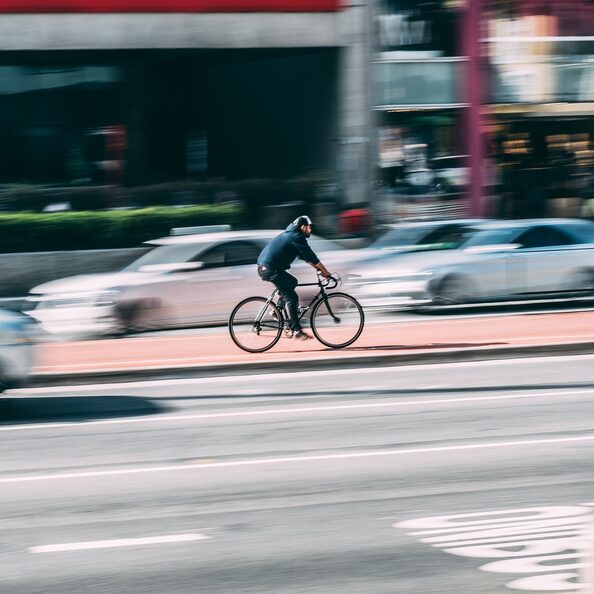 Ein Mensch auf einem Fahrrad fährt mitten auf der Straße entgegen dem Autoverkehr auf einem Radweg. Die Autos sind alle unschraf, der Radfahrer ist im Zentrum des Bildes und scharf