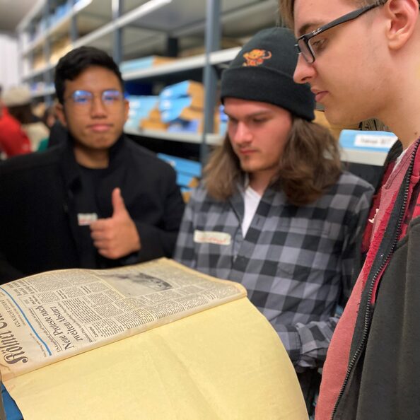 Mehrere junge Männer schauen auf ein alte Zeitung im Archiv, einer macht ein "Daumen hoch"-Zeichen