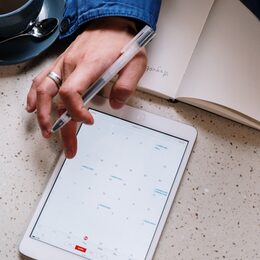eine Hand mit Kugelschreiber einen digitalen Kalender am Tablet mit einem Papier/Buchkalender ab