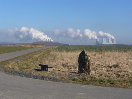 Eine Straßenkreuzung mit Steinbank und Megalith auf einer Wiese. Im Hintergrund Industrieanlagen mit viel Wolkenbildung
