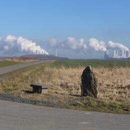 Eine Straßenkreuzung mit Steinbank und Megalith auf einer Wiese. Im Hintergrund Industrieanlagen mit viel Wolkenbildung