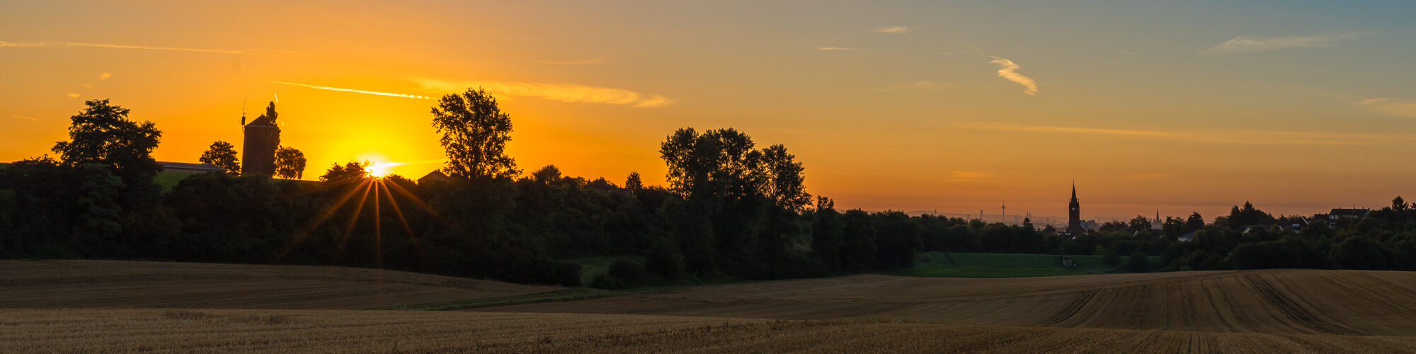 Sonnenaufgang über einem abgeernteten Getreidefeld, links die Sonne neben einer alten Mühle, rechts am Horizont winzig die Kölner Skyline mit Colonius und Kölner Dom