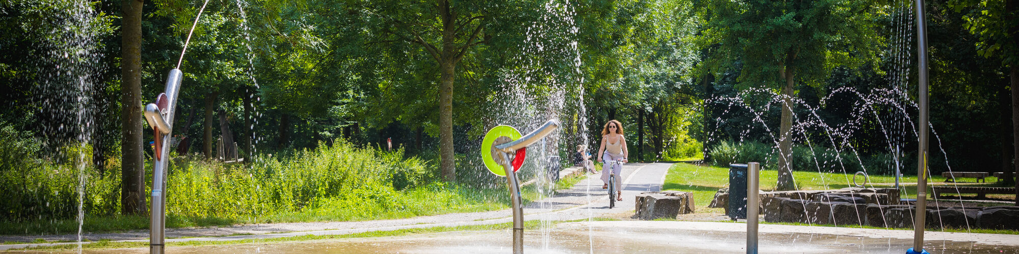 Grüne Parklandschaft, vorne ein Wasserspielplatz mit mehreren Rohren und Wasserfiguren. Von hinten kommt eine Frau mit dem Fahrrad zentral ins Bild